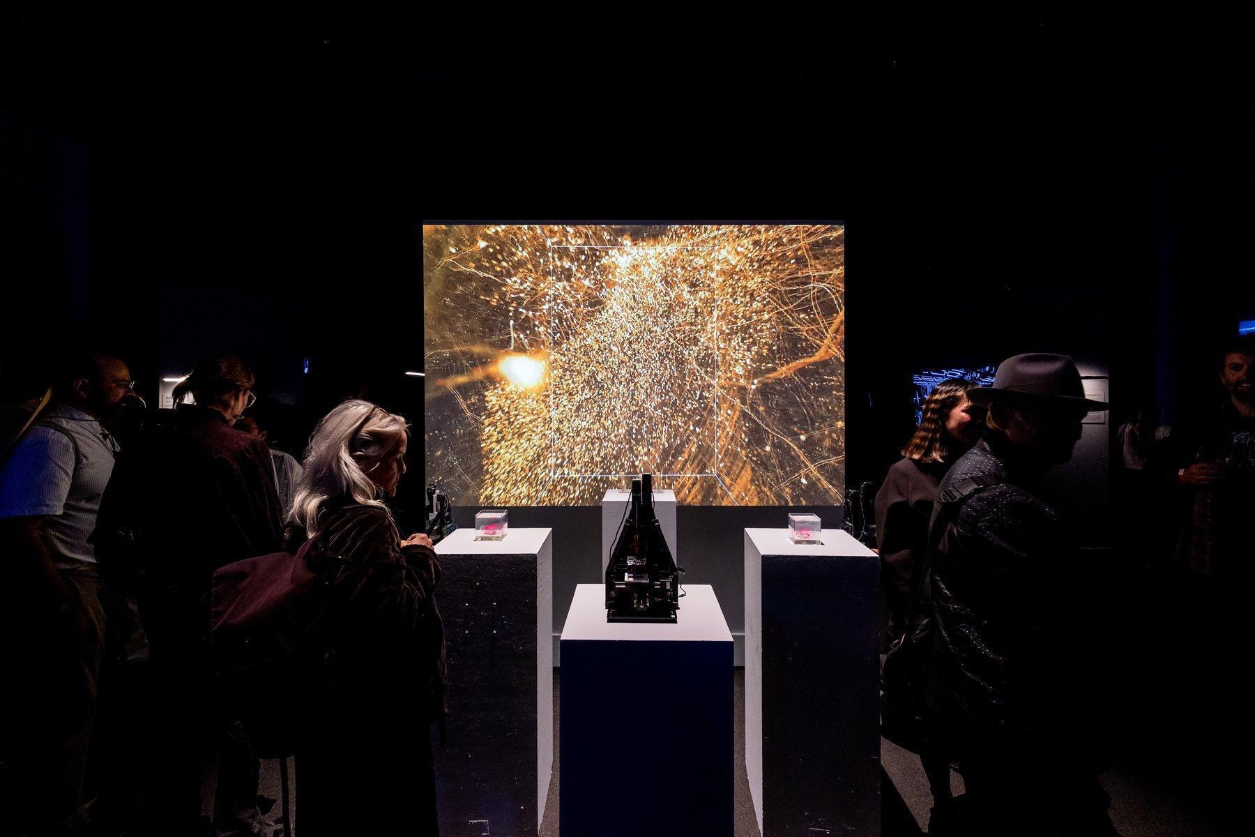 A dark gallery crowded with visitors; three white plinths with small cubes and a bioprinter stand before a large projection of a golden network.