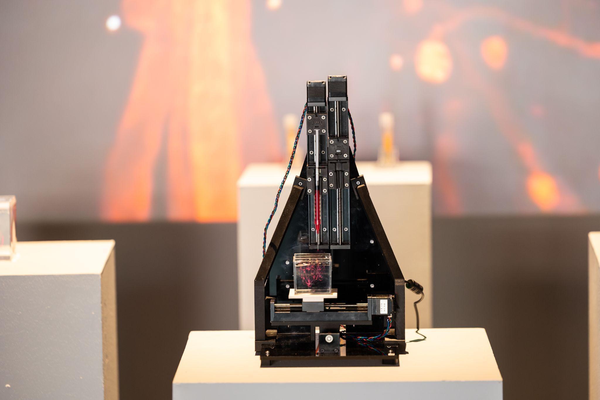 A compact black A-frame bioprinter on a white plinth printing into a small cube, with orange light blurred behind it.