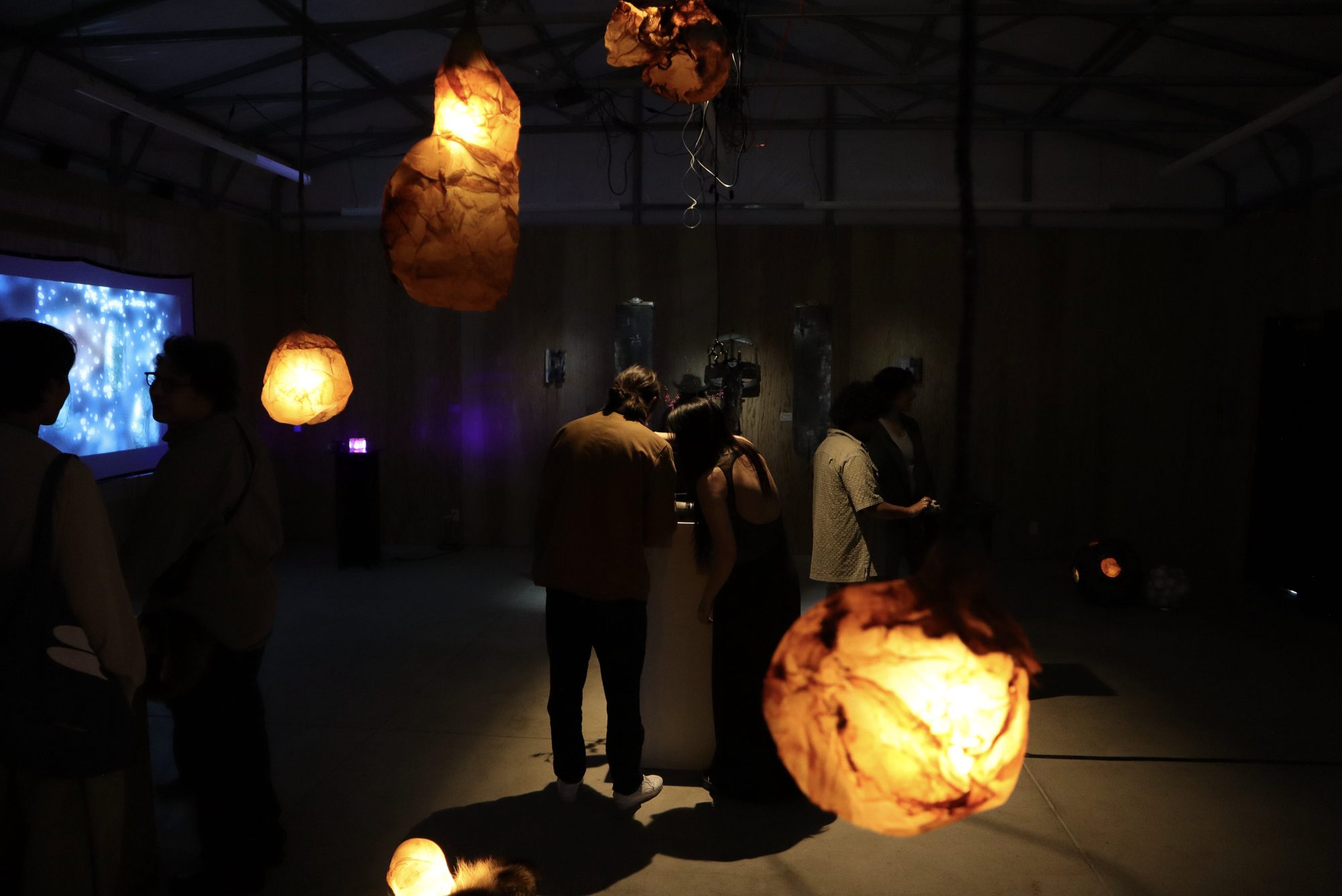 A dark gallery with glowing, rock-like amber lamps hanging and resting on the floor among visitors, a blue projection at the far wall.