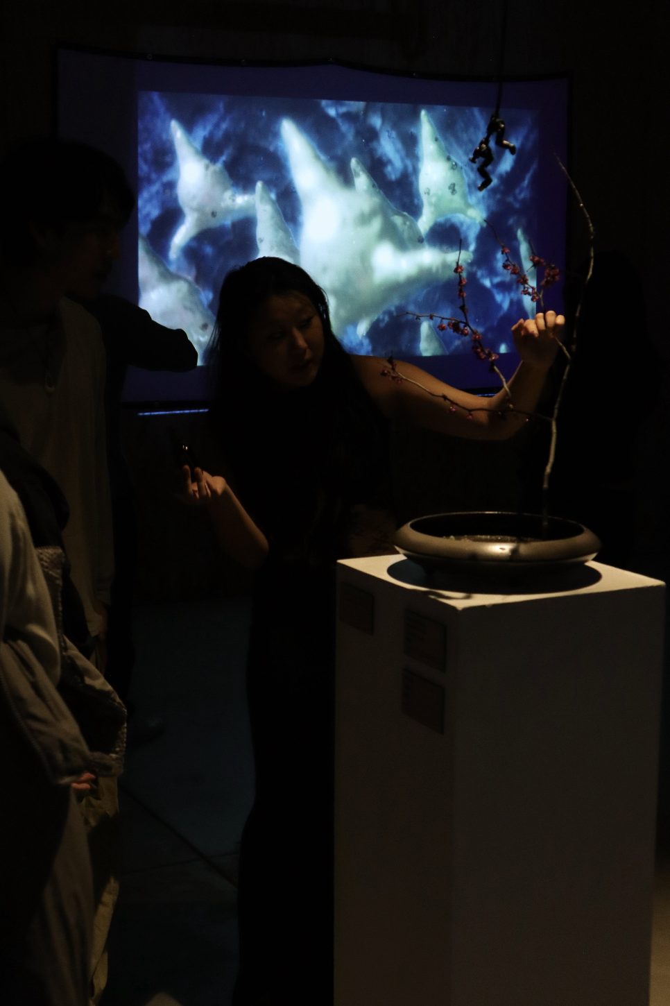 A visitor beside Synthetic Cesia on its plinth, in front of a large projection of blue, cloudlike forms.