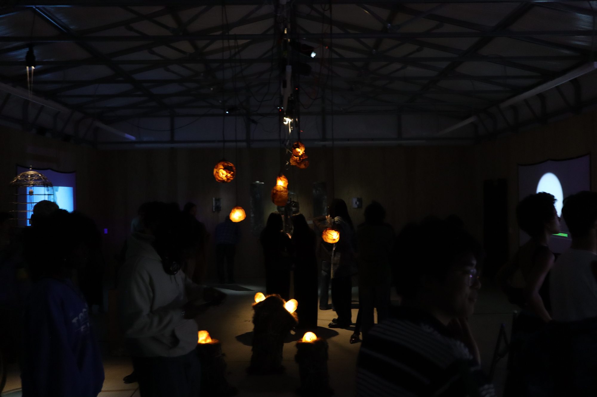A wide view of the gallery in near darkness, small glowing works on the floor and visitors throughout.