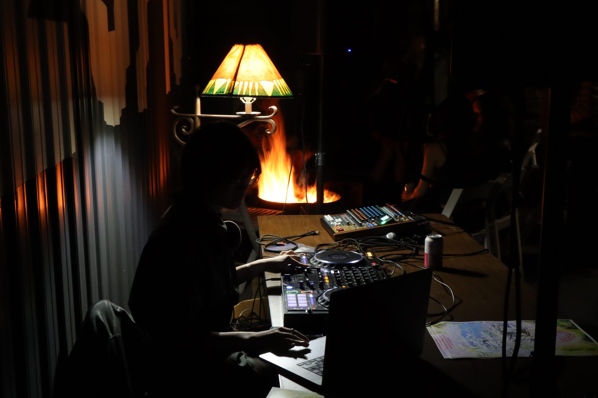 A performer at a mixing desk beside a lit table lamp and a small flame, in the dark gallery.
