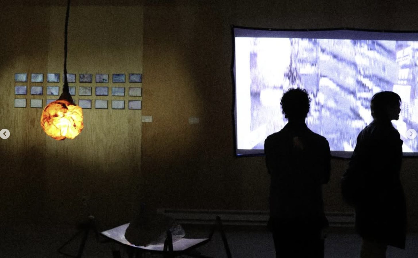 An amber lamp hanging in front of a projection, two visitors silhouetted, a row of small framed images on the wall.