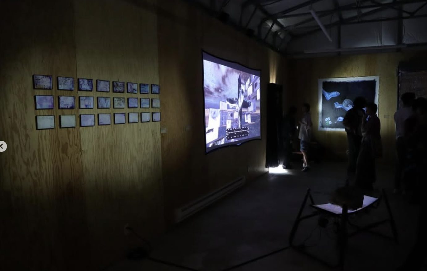 A long view of the gallery: projections on two walls, a row of small framed images, a lamp on the floor.