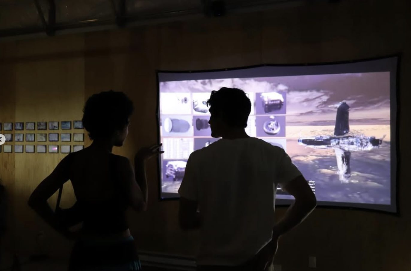 Two visitors silhouetted against a large projection of a pale, airy landscape.